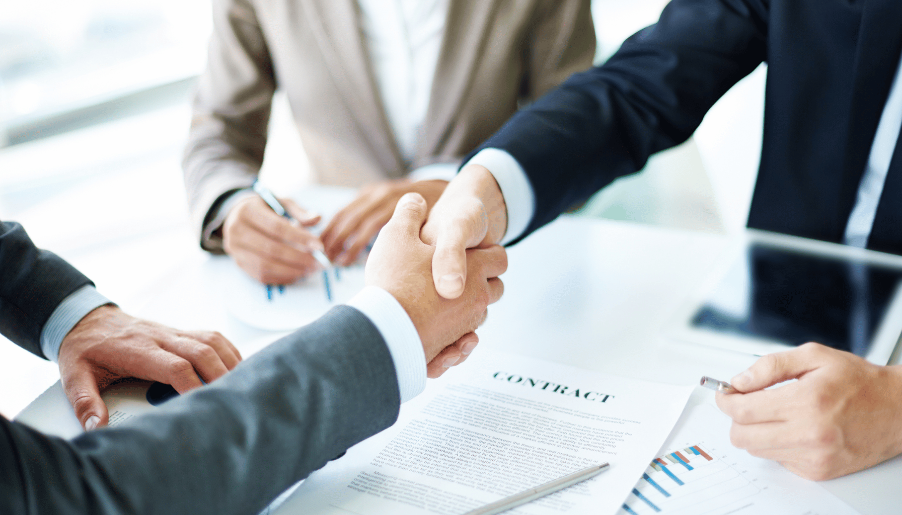 Two people shaking hands over a contract with others observing.