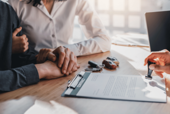 Two people at a desk with a contract, a laptop, and car keys, suggesting a vehicle purchase.