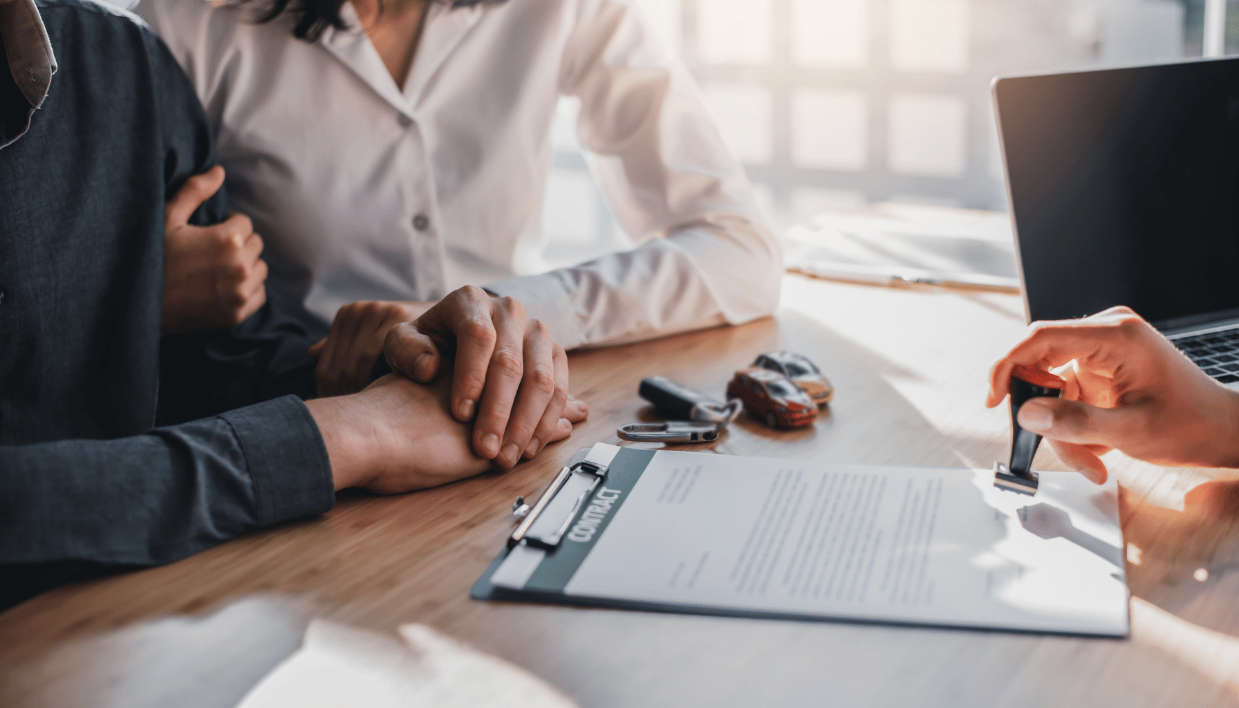 Two people at a desk with a contract, a laptop, and car keys, suggesting a vehicle purchase.