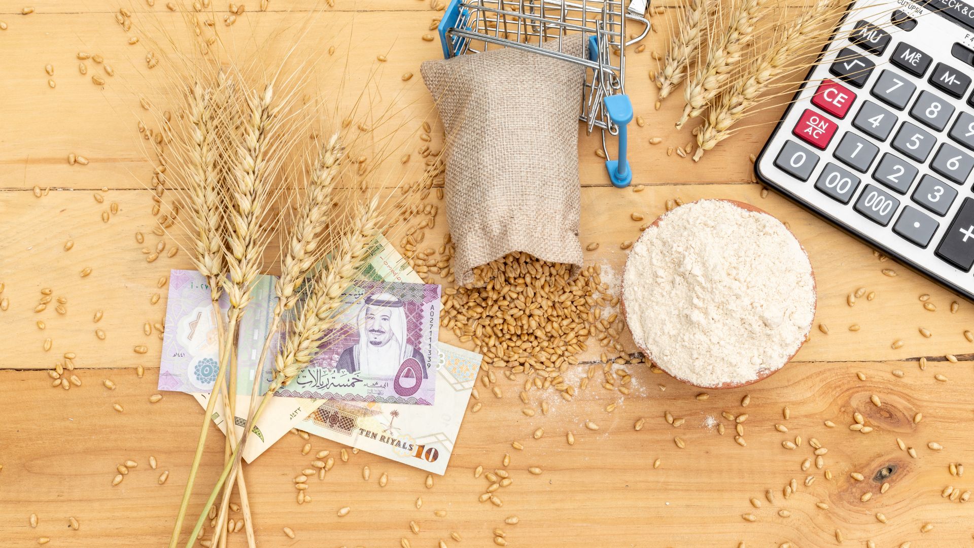 A wooden table displays wheat, a stack of money, and a calculator, symbolizing agriculture and finance.